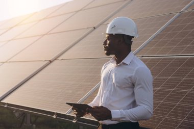 Sert şapkalı Afrikalı Amerikalı mühendis elinde dijital tablet tutarken dışarıda durup güneş panellerini inceliyor. Güneş bu yenilenebilir enerji sahnesi için doğal ışıklandırma sağlıyor..