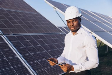 Beyaz şapkalı profesyonel bir Afrikalı Amerikalı mühendis açık mavi gökyüzünün altında güneş panellerinin önünde elinde bir tablet tutuyor. Sahne yenilenebilir enerji teknolojisini temsil ediyor..