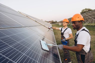 Two skilled workers wearing helmets and gloves are cleaning solar panels in a green outdoor environment. This image captures a scene of teamwork, sustainability, and renewable energy maintenance. clipart