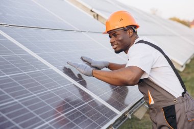 Turuncu bir şapka takan Afrikalı Amerikalı teknisyen ve eldivenler açık hava güneş çiftliğinde güneş panellerini inceliyor. Görüntü yenilenebilir enerji kavramlarını ve profesyonel bakımı yakalar.