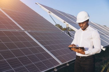 Sert şapka takan profesyonel bir Afrikalı Amerikalı mühendis güneş panellerini gün doğumunda incelemek için bir tablet kullanır. Görüntü, yenilenebilir enerji ve güneş endüstrisindeki teknolojiyi iletiyor..