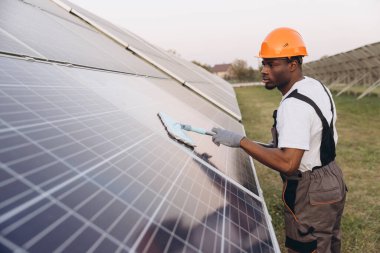 Sert şapkalı ve tulumlu bir işçi dışarıda güneş panellerini temizler ve ideal enerji verimliliğini sağlar. Görüntü yenilenebilir enerji bakımı ve çevresel sorumluluğu vurguluyor.