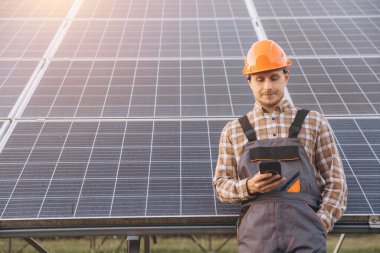 Başlık takan ve tulumlu bir mühendis güneş panellerinin yanında dururken akıllı telefonunu kontrol ediyor. Sahne, yenilenebilir enerji endüstrisindeki sürdürülebilir enerji uygulamalarını ve modern teknolojiyi yakalıyor..