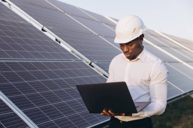 Sert şapka takan Afrikalı Amerikalı mühendis güneş panellerini incelemek için bir dizüstü bilgisayar kullanıyor. Görüntü yenilenebilir enerji, teknoloji ve çevresel sürdürülebilirliği temsil ediyor.