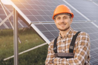 Genç bir mühendis, güneş paneli alanında güvenlik donanımı giyerken, yenilenebilir enerji ve sürdürülebilirlik sergileyerek güvenle gülümsüyor. Batan güneş bu çevre dostu manzaraya sıcaklık katıyor..