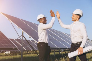 Güneş panelleri önünde güvenlik miğferleri takan iki mühendis yenilenebilir enerjideki başarıyı ve takım çalışmasını kutluyor. Sahne yenilikçi işbirliğini ve sürdürülebilir enerji çözümlerini temsil ediyor