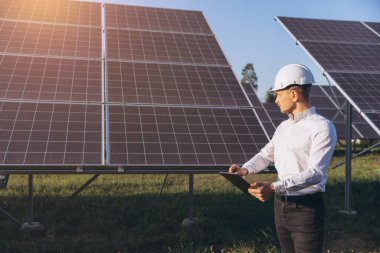 Beyaz kasklı bir mühendis güneş panellerini bir güneş çiftliğinde inceliyor. Görüntü teknolojiyi, yenilenebilir enerjiyi ve sürdürülebilirliği vurguluyor, çevre dostu enerjiye olan bağlılığını gösteriyor.