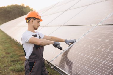 Güvenlik kasklı bir teknisyen yenilenebilir enerji ve sürdürülebilirliği simgeleyen bir matkapla güneş panellerini inceliyor. Ayarlar temiz enerji ve çevresel bilinç üzerine odaklanmayı öneriyor..