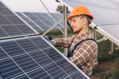 Sert şapkalı erkek mühendis bir güneş çiftliğindeki güneş panellerini inceleyerek verimli enerji üretimini sağlıyor. Bu resim yenilenebilir enerji teknolojisini ve sürdürülebilir gelişimi vurgular.