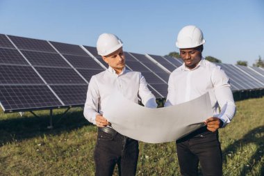 Güneş panelleriyle çevrili bir güneş enerjisi çiftliğinin planlarını inceleyen iki mühendis. Görüntü yenilenebilir enerji kavramları ve takım çalışması iletir.