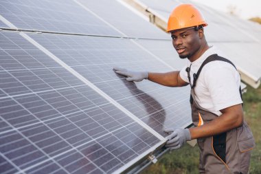 Sert şapkalı Afrikalı Amerikalı mühendis güneş panellerini kontrol ediyor, yenilenebilir enerji ve sürdürülebilirliği vurguluyor. Odaklanmış ifadesi temiz enerji teknolojisinin önemini vurguluyor..