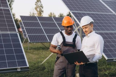 Two engineers collaborate onsite amidst a solar panel array, evaluating data on a laptop. They wear safety helmets, emphasizing renewable energy solutions and teamwork in the green energy sector. clipart