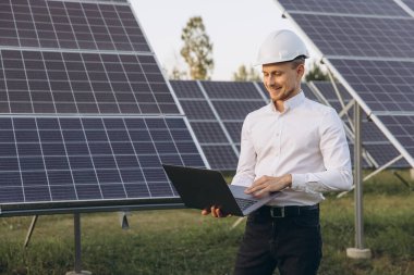 Güneş panelleri alanında dizüstü bilgisayar kullanan beyaz başlıklı bir mühendis yenilenebilir enerji ve teknoloji sergiliyor. Temiz enerji çözümlerinde sürdürülebilir uygulamalar ve yenilikler sunar.