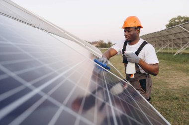 Güvenlik donanımlı Afrikalı Amerikalı erkek işçi güneş panellerini bir güneş çiftliğinde temizliyor. Bakım için bez ve sprey kullanıyor, verimli güneş enerjisi üretimi sağlıyor..