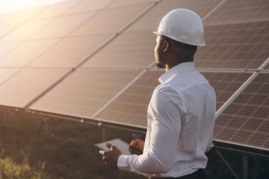 Günbatımında yenilenebilir enerji alanındaki güneş panellerini incelerken başlık takan Afrikalı Amerikalı mühendis. Güneş enerjisindeki sürdürülebilirlik, temiz enerji ve teknolojik gelişmeyi vurguluyor.