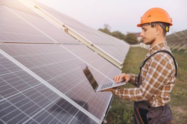 Sert şapka takan erkek bir mühendis, güneş panellerini kontrol etmek için bir dizüstü bilgisayar kullanıyor. Yenilenebilir enerji ve teknolojik gelişmeleri destekliyor..