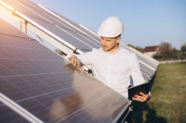 Sert şapka takan bir mühendis güneş panellerini güneşli bir alanda inceleyerek sürdürülebilir enerjiyi destekliyor. Yenilenebilir enerji ve teknoloji kavramları için mükemmel. Çevre dostu iş kullanımı için ideal.