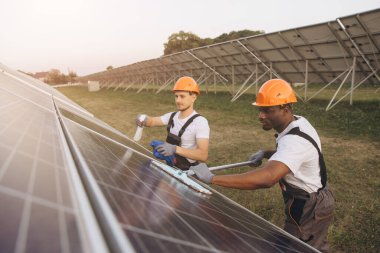 Two technicians in orange helmets clean solar panels at a solar farm. They demonstrate teamwork and maintenance in renewable energy under the warm glow of the setting sun. clipart