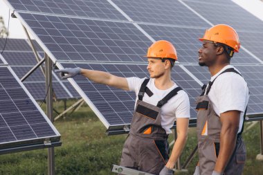 Sert şapkalı ve üniformalı iki mühendis sürdürülebilir bir enerji projesi için güneş panellerini inceliyor. Güneş çiftliği ortamında kurulum sürecini tartışıp analiz ediyorlar..