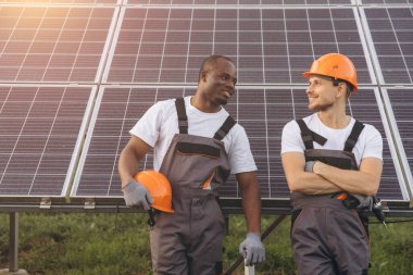 Güvenlik teçhizatı kullanan iki mühendis, güneş panelleri önünde güvenli bir şekilde duruyor, güneşli bir ortamda takım çalışmasını ve sürdürülebilir enerji çözümlerini somutlaştırıyorlar. Yenilik ve işbirliği iletiyorlar.