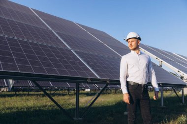 Sert şapka takan bir mühendis büyük güneş panellerinin yanında güven içinde duruyor. Görüntü, temiz enerji ve sürdürülebilir teknolojinin görüntüsünü açık mavi gökyüzü altında yakalıyor..