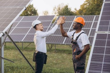 Sert şapka takan iki mühendis güneş panellerinin yanında beşlik çakarak kutluyor. Bu resim, sürdürülebilir uygulamaları teşvik ederek, açık hava ortamında takım çalışması ve yenilenebilir enerji çözümleri yakalar.