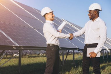 Güneş enerjisi çiftliğinde iki iş adamı el sıkışıyor. Bunlar takım çalışmasını ve sürdürülebilir iş uygulamalarını temsil ediyor. Arka planda açık mavi gökyüzünün altında güneş panelleri bulunuyor..