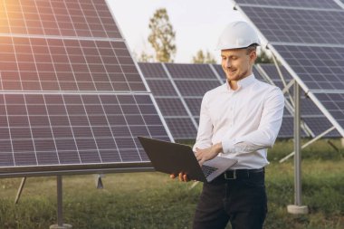 Beyaz şapkalı kendinden emin bir mühendis güneş panellerini güneşli bir alanda dizüstü bilgisayar kullanarak inceleyerek sürdürülebilir bir çevrede yenilenebilir enerji teknolojisi ve yenilik sergiliyor..