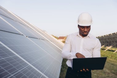 Sert şapkalı Afrikalı Amerikalı mühendis yenilenebilir enerji ve teknolojiyi sembolize eden bir dizüstü bilgisayarla güneş panellerini inceliyor. Bu görüntü sürdürülebilir mühendisliği, temiz enerjiyi ve yeniliği temsil eder..
