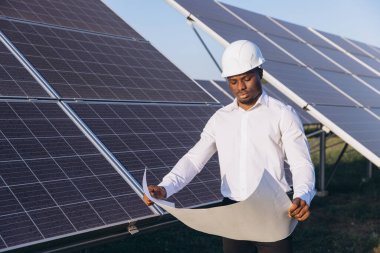 Beyaz şapkalı Afrikalı Amerikalı mühendis güneş panellerinin yanındaki planları inceliyor. Görüntü, temiz enerji ortamında odak, takım çalışması ve yenilenebilir enerji yeniliklerini yakalar.