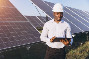 Beyaz kasklı Afrika kökenli Amerikalı bir erkek mühendis güneş panellerini kontrol ederken bir tablet kullanıyor. Görüntü, profesyonel bir çalışmada yenilenebilir enerji, teknoloji ve sürdürülebilirliği vurguluyor.