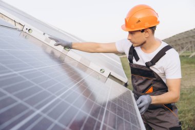 Kask takan genç bir mühendis dışarıda güneş panellerini inceliyor. Görüntü, yenilenebilir enerji uygulamaları ve sürdürülebilirlik çabalarını yakalıyor. Temiz enerji ve mühendislik konuları için ideal.