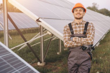 Güvenlik teçhizatı takan gülümseyen bir mühendis güneşli bir alanda güneş panellerinin yanında, yenilenebilir enerji ve sürdürülebilir mühendislik çözümlerini vurguluyor. Görüntü iyimserliği iletir.