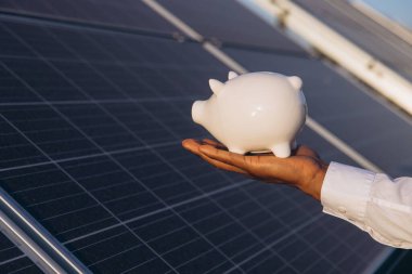 Bir el, tasarruf ve yenilenebilir enerji yatırımını sembolize eden güneş panelleri önünde beyaz bir kumbara tutuyor. Görüntü finansal güvenlik ve sürdürülebilir enerji çözümleri sunar.