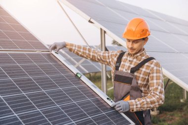 Sert şapka takan yetenekli bir mühendis güneş panellerini bir güneş çiftliğinde inceliyor. Görüntü yenilenebilir enerji teknolojileri ve çevresel sürdürülebilirlik üzerine odaklanmayı betimliyor.