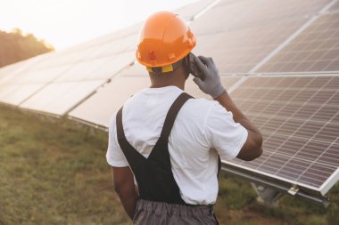 Güvenlik teçhizatı takan bir mühendis cep telefonuyla konuşurken güneş panellerini kontrol ediyor. Görüntü yenilenebilir enerji, teknoloji ve güneş enerjisi alanındaki iletişim temalarını yakalar.