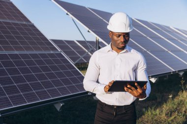 Siyah şapkalı Afrikalı Amerikalı mühendis güneş panellerini yönetmek için dijital tablet kullanıyor. Güneş çiftliği ortamında yenilenebilir enerji, teknoloji ve sürdürülebilir kalkınma kavramı.