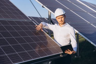 Yenilenebilir enerji çiftliğindeki güneş panellerini inceleyen ve veri analizi için tablet tutan bir başlık mühendisi. Güneş enerjisi projelerinde yenilik, sürdürülebilir enerji ve modern teknolojiyi vurguluyor.