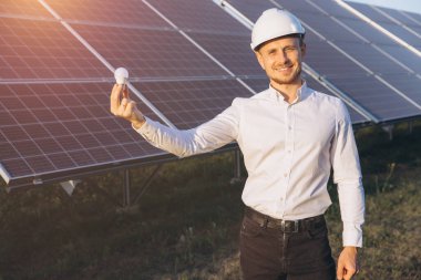 Sert şapka takan gülümseyen bir mühendis güneş panelleri önünde yenilenebilir enerji ve yeniliği simgeleyen bir LED ampulü tutuyor. Güneş ışığı sürdürülebilir enerji kavramını geliştirir.