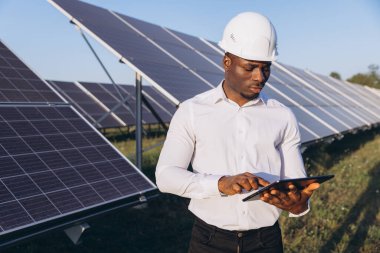 Başlık takan Afrikalı Amerikalı mühendis güneş panellerini incelemek için bir tablet kullanıyor. Görüntü yenilenebilir enerji, sürdürülebilirlik ve teknolojik yeniliği yansıtıyor.