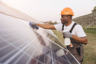 A dedicated African American Male technician wearing safety gear cleans solar panels using cleaning solution and tools. This maintenance work ensures optimal solar energy efficiency. clipart