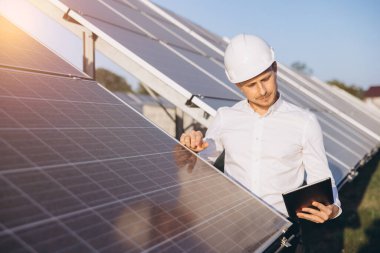 Başlık takan mühendis, dışarıdaki güneş panellerini incelerken dijital tablet kullanıyor. Görüntü yenilenebilir enerji, teknoloji ve sürdürülebilirlik kavramlarını iletir.