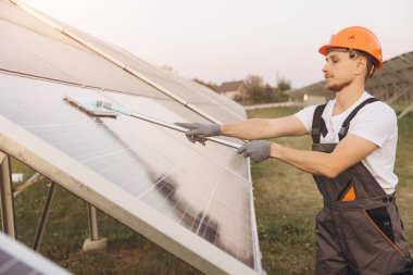 İş kıyafetli ve şapkalı bir adam güneş panellerini fırçayla temizliyor, kırsal alanda yenilenebilir enerji bakımı ve sürdürülebilirlik çabalarını temsil ediyor..