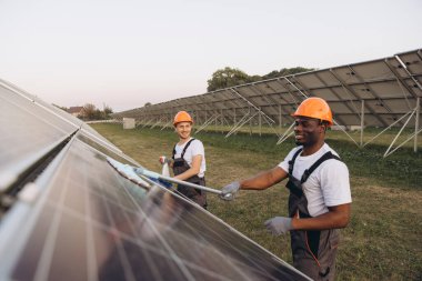 Two workers in orange helmets clean solar panels outdoors. They are smiling and wearing protective gear, promoting renewable energy and teamwork in a sustainable environment. clipart