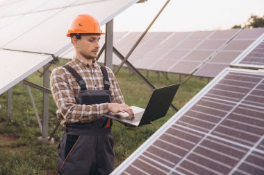 Güneş panellerinin güneş çiftliğindeki işleyişini izlemek için dizüstü bilgisayar kullanan koruyucu ekipman mühendisi. Görüntü yenilenebilir enerji teknolojisi, sürdürülebilirlik ve modern mühendislik uygulamalarını temsil etmektedir..