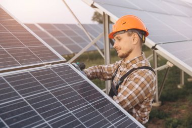 Sert şapkalı bir erkek mühendis, yenilenebilir enerji ve sürdürülebilirliği vurgulayarak açık hava ortamında güneş panellerini inceliyor. Görüntü profesyonelliği ve çevresel sorumluluğu aktarıyor.
