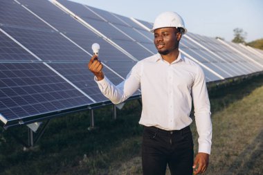Beyaz gömlekli ve kasklı Afro-Amerikan mühendis güneş panellerinin önünde dururken elinde ampul tutuyordu. Görüntü yenilenebilir enerji ve sürdürülebilir teknolojiyi temsil ediyor..