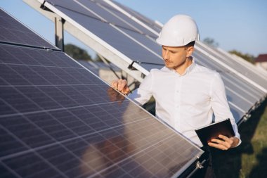 Odaklanmış bir mühendis, yenilenebilir enerji ve sürdürülebilir teknolojiyi simgeleyen açık mavi gökyüzünün altındaki güneş panellerini inceliyor. Sahne yenilikleri ve temiz enerji ilerlemelerini aktarıyor..