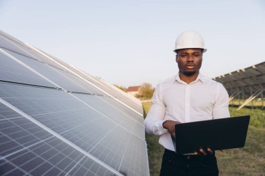 Sert şapkalı Afrikalı Amerikalı mühendis güneş panellerini dizüstü bilgisayarla inceliyor. Güneşli bir ortamda yenilenebilir enerji ve sürdürülebilir teknolojiyi vurguluyor..