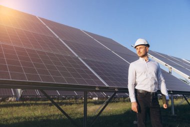 Sert şapkalı bir mühendis güneş panellerini açık bir gökyüzünün altında kontrol ediyor. Güneş ışığı modern teknolojiyi ve yenilenebilir enerjiyi vurguluyor. Sürdürülebilirlik ve yenilik kavramları için ideal.